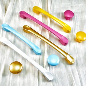 May include: A selection of colourful cabinet hardware, including drawer pulls and knobs. The pulls are in white, light blue, pink, and gold, while the knobs are in gold, pink, yellow, and light blue. The hardware is arranged on a white, textured surface.