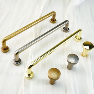 May include: An assortment of cabinet hardware, including three bar pulls in gold, silver, and brass tones, and three matching round knobs. The hardware is displayed on a white, textured surface.