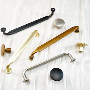 May include: A collection of cabinet hardware in various finishes, including black, brass, and silver. The assortment features knobs and pulls in different shapes and sizes, suitable for furniture or cabinetry. The hardware is displayed on a white surface.