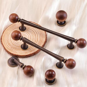 Op de afbeelding: Een verzameling houten meubelbeslag, waaronder twee lange handgrepen en twee knoppen, met een donker bronzen afwerking. De handgrepen hebben een cilindrische staaf met een houten bol aan elk uiteinde. De knoppen hebben een houten bol op een bronzen voet.