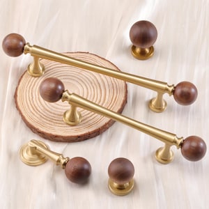 May include: A collection of cabinet hardware featuring brass-toned pulls and knobs with dark brown wooden ball accents. The pulls are long and slender, while the knobs are spherical. The hardware is displayed on a wooden slice.