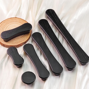 May include: A collection of black wooden cabinet hardware, including various sizes of pulls and knobs. The hardware is displayed on a white fabric background, with a small wooden slice in the background.