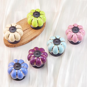 Pode incluir: Uma coleção de puxadores de armário de cerâmica coloridos em várias tonalidades, incluindo verde, creme, rosa, azul e roxo. Cada puxador tem um design floral com detalhes dourados e um centro escuro. Alguns puxadores estão dispostos numa tábua de madeira.