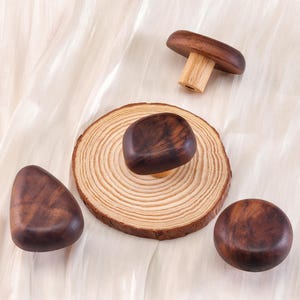 Może przedstawiać: Drewniane przedmioty w kształcie grzyb&oacute;w o r&oacute;żnych kształtach. Jeden to kompletny grzyb, drugi to kapelusz grzyba na drewnianej podstawie, a pozostałe to gładkie, zaokrąglone kształty. Obiekty wykonane są z ciemnobrązowego drewna.