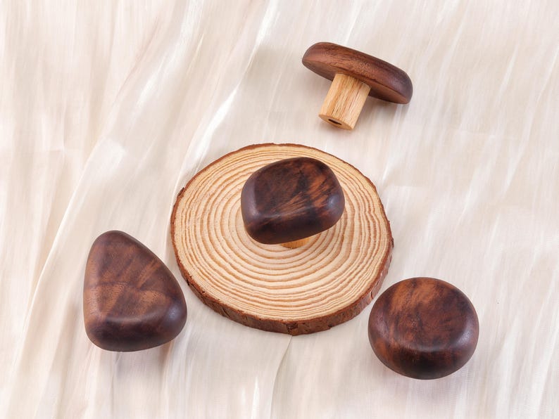 Może przedstawiać: Drewniane przedmioty w kształcie grzyb&oacute;w o r&oacute;żnych kształtach. Jeden to kompletny grzyb, drugi to kapelusz grzyba na drewnianej podstawie, a pozostałe to gładkie, zaokrąglone kształty. Obiekty wykonane są z ciemnobrązowego drewna.