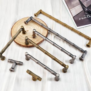 May include: A collection of bamboo-style cabinet pulls in various metallic finishes, including gold, silver, and bronze. The hardware is arranged on a white surface, showcasing the different sizes and designs.