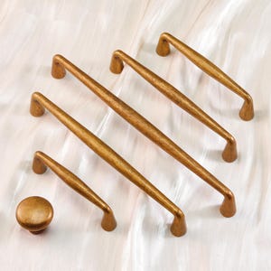 May include: A collection of antique brass cabinet hardware, including several pull handles and a single knob. The handles vary in size, with a curved design and a warm, aged finish. The hardware is arranged on a white surface.
