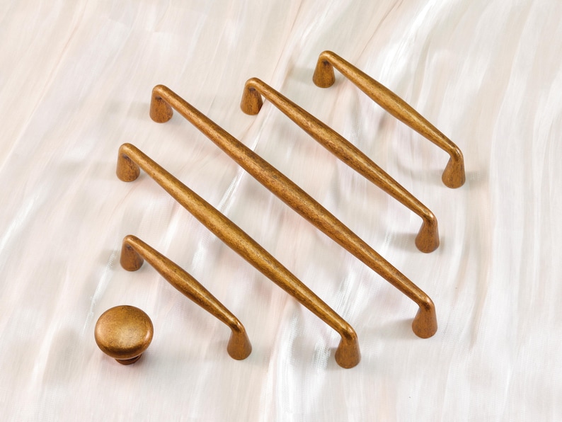 May include: A collection of antique brass cabinet hardware, including several pull handles and a single knob. The handles vary in size, with a curved design and a warm, aged finish. The hardware is arranged on a white surface.
