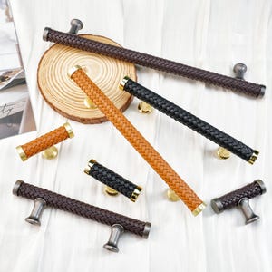 May include: An assortment of cabinet pulls with a woven leather design in various colors: brown, black, and tan. The pulls have brass or silver-toned hardware. The image showcases different sizes and styles of the cabinet hardware.