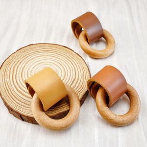 Peut inclure: Anneaux de dentition en bois avec des accents de cuir dans les tons marron et beige, pr&eacute;sent&eacute;s sur une surface blanche. Un anneau est associ&eacute; &agrave; une tranche de bois. Les anneaux sont con&ccedil;us pour les b&eacute;b&eacute;s.