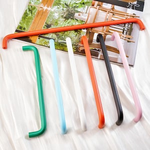 Może przedstawiać: Kolekcja kolorowych, zakrzywionych metalowych uchwytów w różnych odcieniach, w tym zielonym, niebieskim, białym, pomarańczowym, czarnym i różowym. Uchwyty są ułożone na białej powierzchni z magazynem w tle. Uchwyty mają nowoczesny, minimalistyczny design.