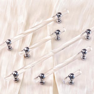 Pode incluir: Um conjunto de puxadores de armário em acrílico transparente com bases cromadas. Os puxadores têm um design cilíndrico torcido e estão dispostos sobre um tecido creme. Ferragens para móveis e decoração de casa.