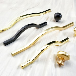 May include: An assortment of cabinet hardware in gold and black. The selection includes curved bar pulls, round knobs, and a mushroom-shaped pull. The hardware is displayed on a white surface, showcasing the variety of designs and finishes.