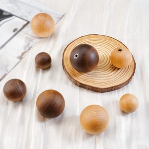Pode incluir: Uma coleção de esferas de madeira em vários tamanhos e tons de castanho e bege. Algumas esferas são castanho escuro, outras bege claro, e uma tem um buraco. Estão dispostas sobre uma superfície branca, com uma fatia de madeira e uma superfície refletora no fundo.