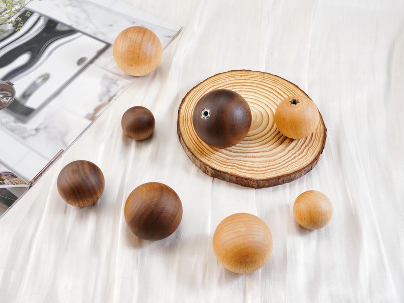 Puede incluir: Una colecci&oacute;n de esferas de madera en varios tama&ntilde;os y tonos de marr&oacute;n y beige. Algunas esferas son de color marr&oacute;n oscuro, otras de color beige claro, y una tiene un agujero. Est&aacute;n dispuestas sobre una superficie blanca, con una rodaja de madera y una superficie reflectante en el fondo.