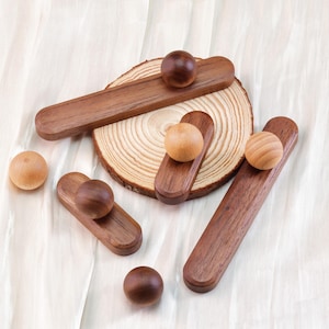 May include: Wooden decorative objects, including small, dark brown wooden balls and oblong wooden pieces. Some pieces have a ball attached. A wooden tree slice is also present. The objects are arranged on a white, textured surface.