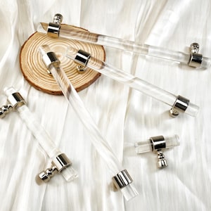 May include: Clear acrylic cabinet pulls with silver-toned hardware. The pulls are cylindrical and have a modern design. They are arranged on a white fabric surface with a wooden slice.