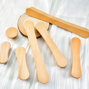 May include: A collection of various wooden craft supplies, including long, flat sticks, rounded paddles, and a small wooden disc. The items are arranged on a white, textured surface, showcasing their natural wood grain and light brown color.