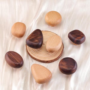 Może przedstawiać: Asortyment drewnianych gałek do szafek w r&oacute;żnych kształtach i odcieniach brązu. Niekt&oacute;re gałki są jasne, inne ciemniejsze i bogatsze w brąz. Gałki ułożone są na białej, teksturowanej powierzchni.