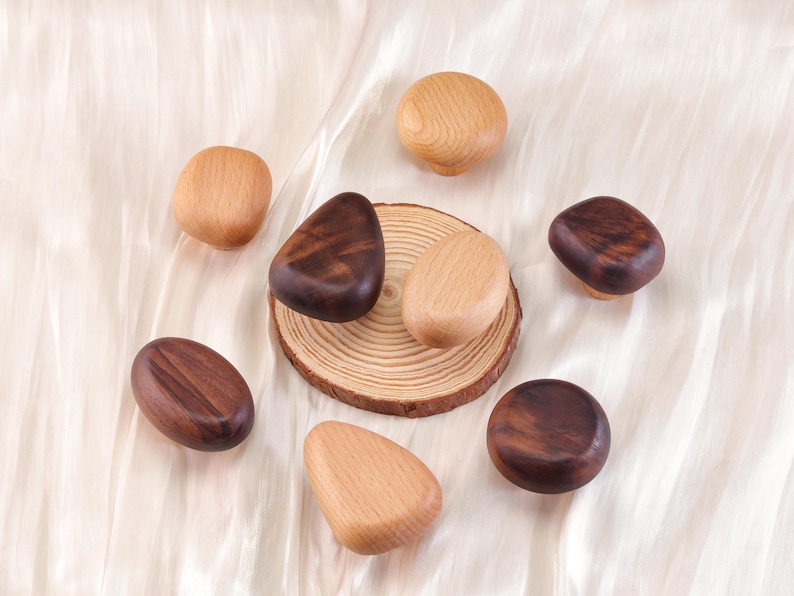 Może przedstawiać: Asortyment drewnianych gałek do szafek w r&oacute;żnych kształtach i odcieniach brązu. Niekt&oacute;re gałki są jasne, inne ciemniejsze i bogatsze w brąz. Gałki ułożone są na białej, teksturowanej powierzchni.