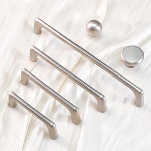 May include: A collection of silver-toned cabinet hardware, including four different-sized bar pulls, a round knob, and a spherical knob. The hardware is arranged on a white, textured surface.