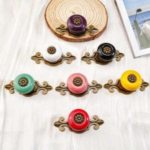 May include: A selection of colourful ceramic cabinet knobs with brass-toned decorative backplates. The knobs are in various colours, including purple, white, black, green, pink, yellow, and red. Each knob features a floral design.