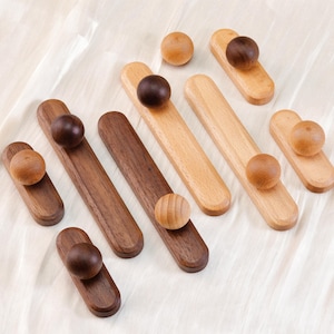 May include: A selection of wooden cabinet pulls and knobs in shades of brown and natural wood. The pulls are rectangular with a rounded knob, while the knobs are spherical. The items are displayed on a white fabric surface.