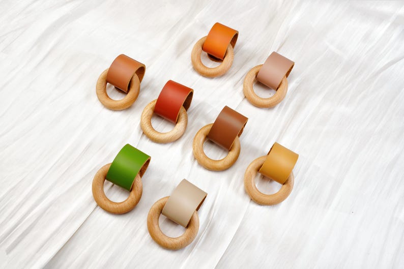Peut inclure: Anneaux de dentition en bois avec des lani&egrave;res en cuir de diff&eacute;rentes couleurs, notamment marron, orange, vert et beige. Les anneaux sont dispos&eacute;s sur une surface blanche textur&eacute;e.