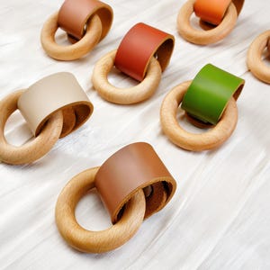 Peut inclure: Anneaux de dentition en bois avec des accents de cuir de diff&eacute;rentes couleurs, dont le marron, le vert et le beige. Les anneaux sont dispos&eacute;s sur une surface blanche, mettant en valeur le grain naturel du bois et les textures du cuir.