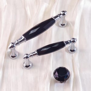 May include: Two cabinet handles and a knob. The handles have a black, faceted center and chrome-colored ends. The knob is a matching black faceted design. All pieces are set against a white, textured background.