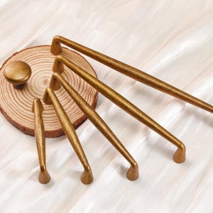 May include: A collection of gold-toned cabinet hardware, including knobs and various sizes of pull handles. The hardware is arranged on a wooden slice, showcasing a range of sizes and designs for home decor and furniture projects.