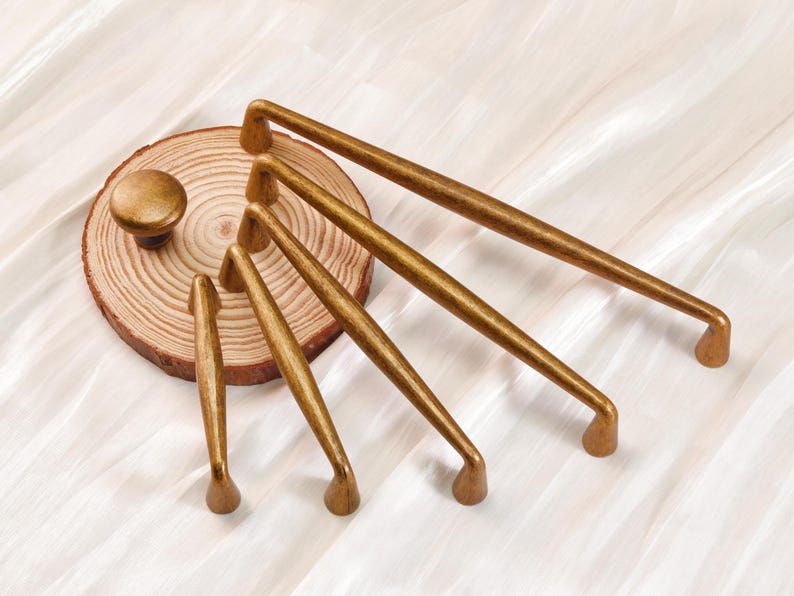 May include: A collection of gold-toned cabinet hardware, including knobs and various sizes of pull handles. The hardware is arranged on a wooden slice, showcasing a range of sizes and designs for home decor and furniture projects.