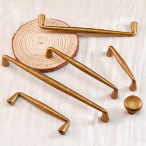 May include: A collection of antique brass cabinet hardware, including various sizes of handles and a knob. The hardware has a warm, aged finish, and is arranged on a light-colored surface with a wooden slice.