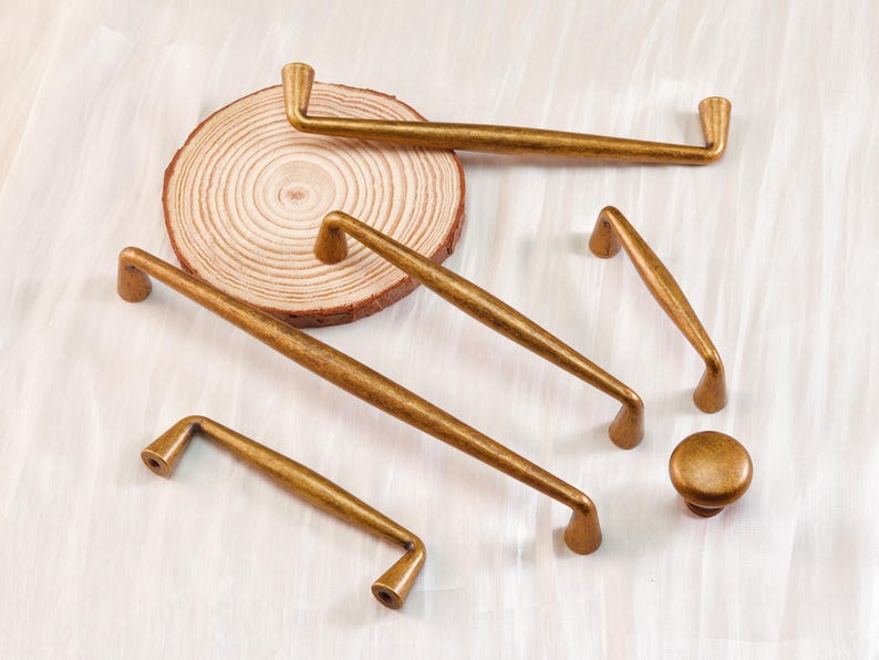 May include: A collection of antique brass cabinet hardware, including various sizes of handles and a knob. The hardware has a warm, aged finish, and is arranged on a light-colored surface with a wooden slice.