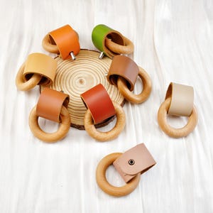 Peut inclure: Divers anneaux de dentition pour b&eacute;b&eacute;s en bois, orn&eacute;s de cuir de diff&eacute;rentes couleurs : orange, vert et marron. Les anneaux sont dispos&eacute;s sur une surface blanche, certains sur une tranche de bois.