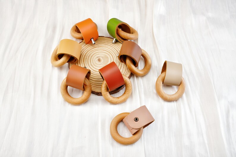 Peut inclure: Divers anneaux de dentition pour b&eacute;b&eacute;s en bois, orn&eacute;s de cuir de diff&eacute;rentes couleurs : orange, vert et marron. Les anneaux sont dispos&eacute;s sur une surface blanche, certains sur une tranche de bois.