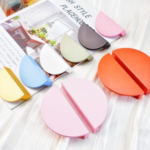 May include: A collection of colorful, semi-circular and circular desk organizers in shades of yellow, blue, white, green, gray, pink, and orange. The organizers are arranged on a magazine and a white, textured surface.