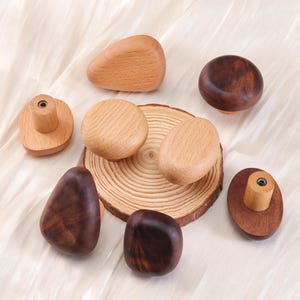 Może przedstawiać: Kolekcja drewnianych gałek do szafek w r&oacute;żnych kształtach i odcieniach drewna. Gałki obejmują kształty zaokrąglone, owalne i nieregularne, niekt&oacute;re z naturalnym usłojeniem drewna, a inne z ciemniejszą bejcą. Ułożone są na jasnej tkaninie.