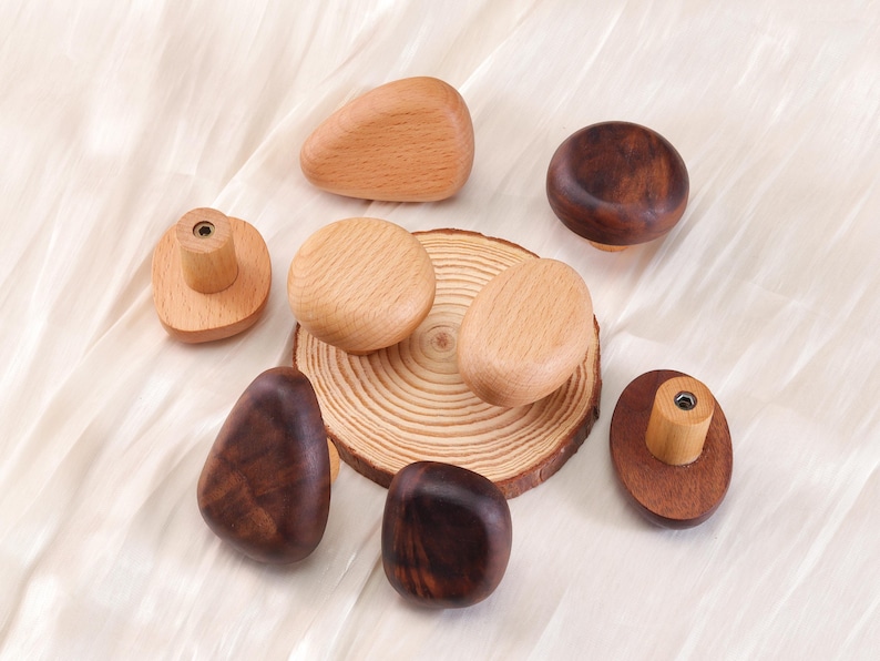 Może przedstawiać: Kolekcja drewnianych gałek do szafek w r&oacute;żnych kształtach i odcieniach drewna. Gałki obejmują kształty zaokrąglone, owalne i nieregularne, niekt&oacute;re z naturalnym usłojeniem drewna, a inne z ciemniejszą bejcą. Ułożone są na jasnej tkaninie.