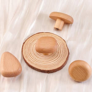 Może przedstawiać: Zestaw drewnianych gałek i uchwyt&oacute;w do szafek w r&oacute;żnych kształtach. Zawiera gałkę w kształcie grzyba, okrągłą gałkę, zaokrągloną tr&oacute;jkątną gałkę i gałkę zamontowaną na plastrze drewna. Gałki wykonane są z jasnego drewna.