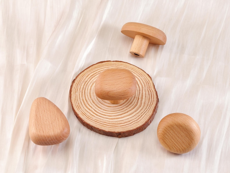 Może przedstawiać: Zestaw drewnianych gałek i uchwyt&oacute;w do szafek w r&oacute;żnych kształtach. Zawiera gałkę w kształcie grzyba, okrągłą gałkę, zaokrągloną tr&oacute;jkątną gałkę i gałkę zamontowaną na plastrze drewna. Gałki wykonane są z jasnego drewna.