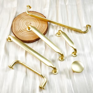 May include: A collection of gold-toned cabinet hardware, including various sizes of handles and a round knob. The hardware is arranged on a light-colored, textured surface, with a wooden slice adding a natural element.