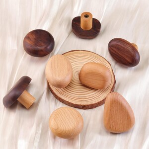 Może przedstawiać: Kolekcja drewnianych gałek i uchwyt&oacute;w do szafek w r&oacute;żnych kształtach i odcieniach drewna. Asortyment obejmuje gałki w kształcie grzyb&oacute;w, owalne uchwyty i okrągłe gałki, wszystkie na neutralnym tle. Gałki wykonane są z naturalnego drewna.