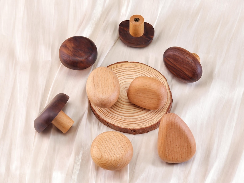 Może przedstawiać: Kolekcja drewnianych gałek i uchwyt&oacute;w do szafek w r&oacute;żnych kształtach i odcieniach drewna. Asortyment obejmuje gałki w kształcie grzyb&oacute;w, owalne uchwyty i okrągłe gałki, wszystkie na neutralnym tle. Gałki wykonane są z naturalnego drewna.