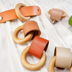 Peut inclure: Ensemble de composants de quincaillerie en cuir et en bois de diff&eacute;rentes couleurs. La quincaillerie comprend des anneaux en bois avec des enveloppes en cuir dans les tons orange, marron, vert et beige. Une pi&egrave;ce a une sangle rectangulaire en cuir avec un anneau en bois.