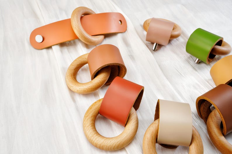 Peut inclure: Ensemble de composants de quincaillerie en cuir et en bois de diff&eacute;rentes couleurs. La quincaillerie comprend des anneaux en bois avec des enveloppes en cuir dans les tons orange, marron, vert et beige. Une pi&egrave;ce a une sangle rectangulaire en cuir avec un anneau en bois.