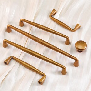 May include: A collection of antique brass cabinet hardware, including several pull handles and a round knob. The handles vary in size and shape, with a slightly curved design. The hardware is displayed on a white, textured surface.