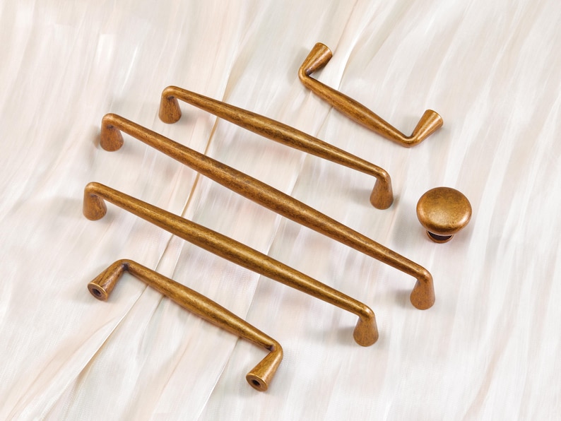 May include: A collection of antique brass cabinet hardware, including several pull handles and a round knob. The handles vary in size and shape, with a slightly curved design. The hardware is displayed on a white, textured surface.