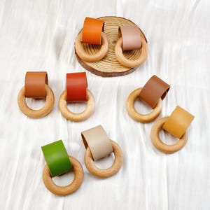 Peut inclure: Anneaux de dentition pour b&eacute;b&eacute; en bois avec des accents en cuir color&eacute; dans les tons orange, marron, rouge, vert, beige et jaune. Les anneaux sont dispos&eacute;s sur une surface blanche, certains sur une petite tranche de bois.