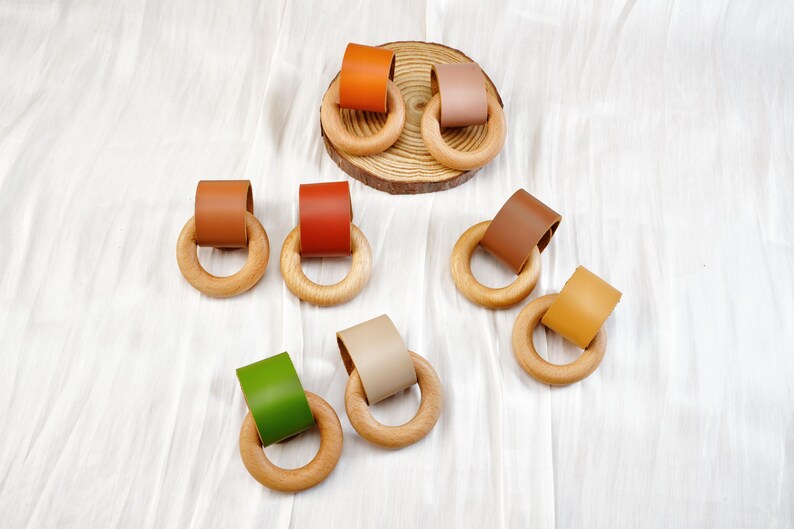 Peut inclure: Anneaux de dentition pour b&eacute;b&eacute; en bois avec des accents en cuir color&eacute; dans les tons orange, marron, rouge, vert, beige et jaune. Les anneaux sont dispos&eacute;s sur une surface blanche, certains sur une petite tranche de bois.